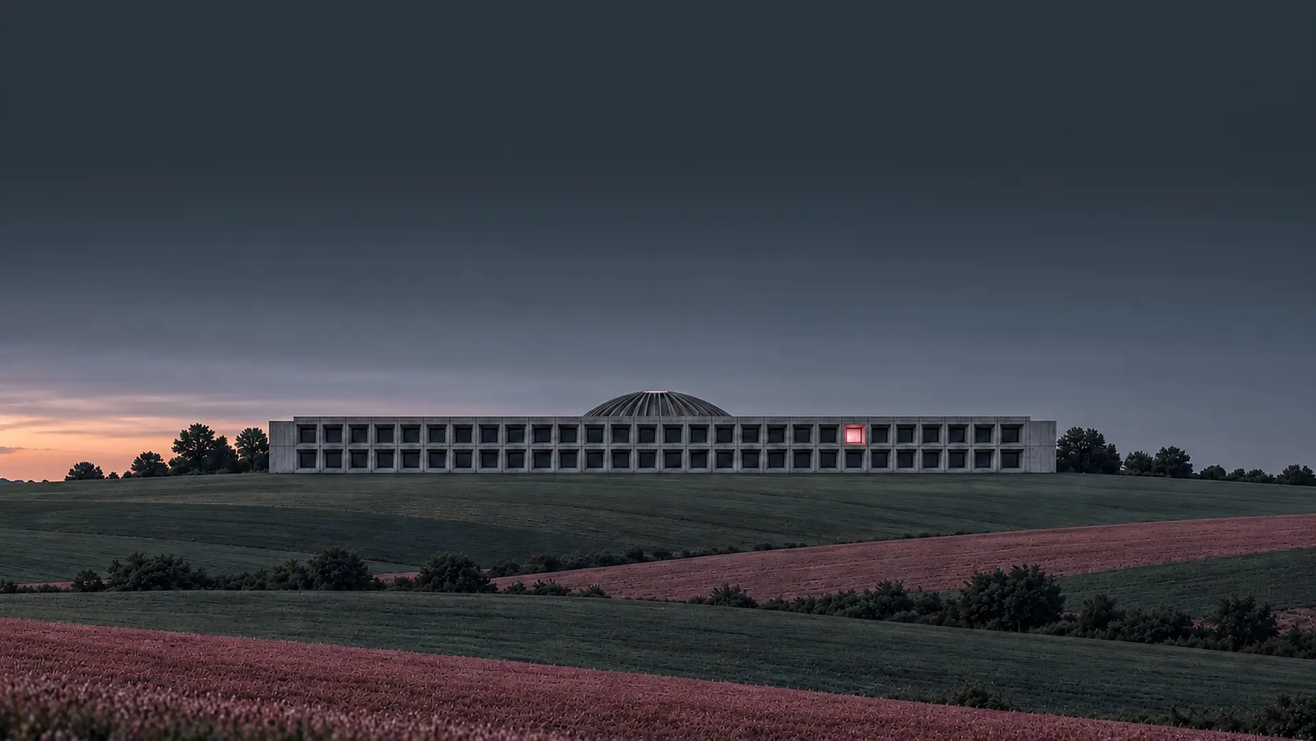 A long, low concrete building on a ridge at dusk, one window lit, banded fields below